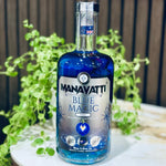 A bottle of Manavatti Blue Magic Vodka with vibrant blue liquid, set on a white marble surface surrounded by green oregano leaves against a dark wooden background.