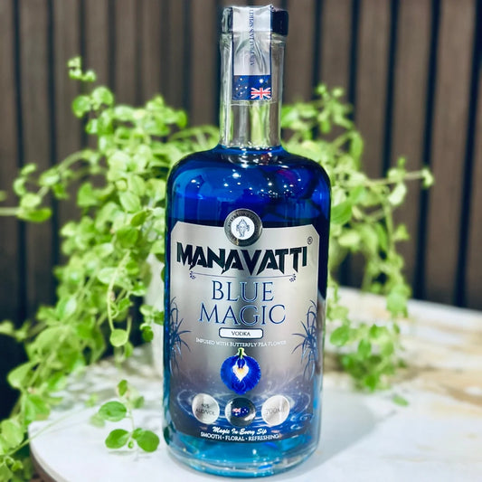 A bottle of Manavatti Blue Magic Vodka with vibrant blue liquid, set on a white marble surface surrounded by green oregano leaves against a dark wooden background.